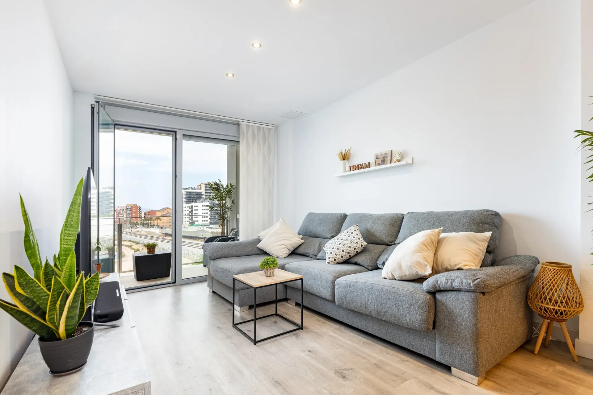 Fotografía inmobiliaria de salón luminoso con sofá gris, balcón y vistas urbanas