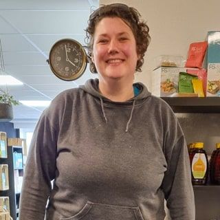 Een lachend persoon met krullend haar en een grijze hoodie staat in een winkel, met op de achtergrond schappen vol producten.