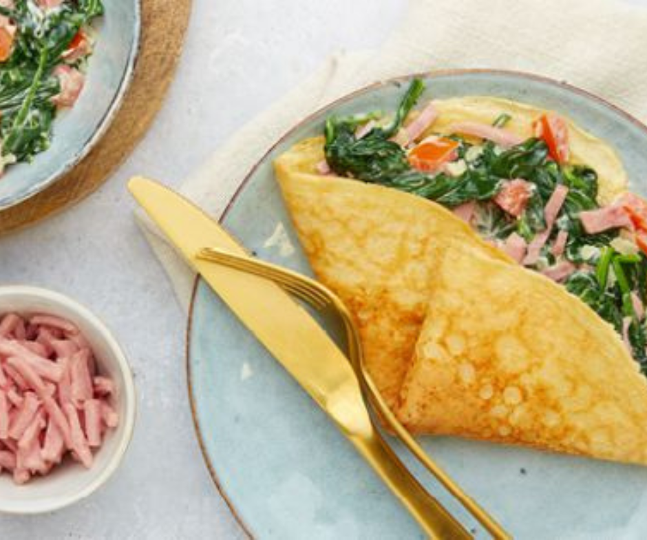 Een gevouwen omelet gevuld met spinazie, blokjes ham en tomaten op een blauw bord, met gouden bestek en een schaaltje ham ernaast.