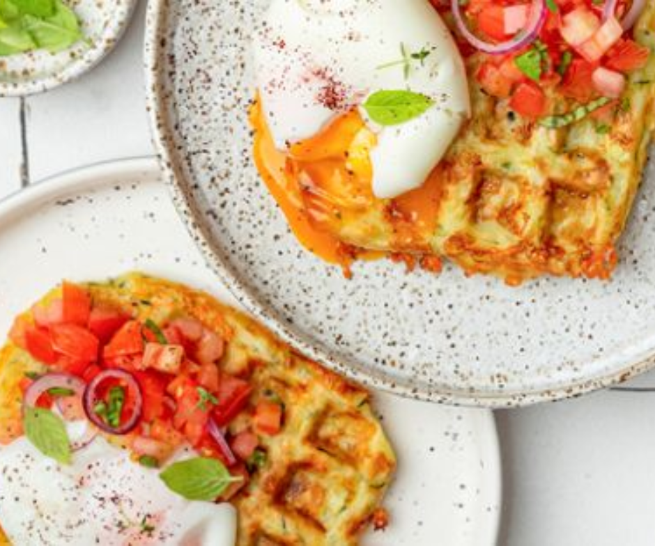 Twee hartige wafels, belegd met gepocheerde eieren, blokjes tomaat, rode ui en verse kruiden, geserveerd op gespikkelde borden.