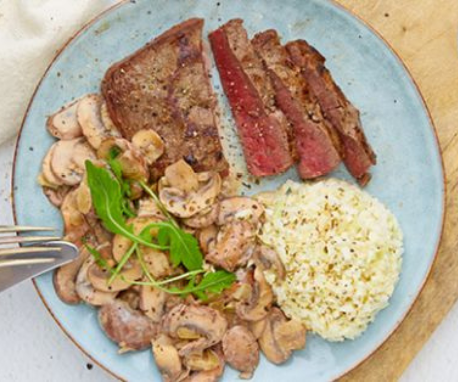 Een gerecht met plakjes medium-rare gebakken biefstuk, romige gebakken champignons met verse groenten en een portie gekruide rijst.