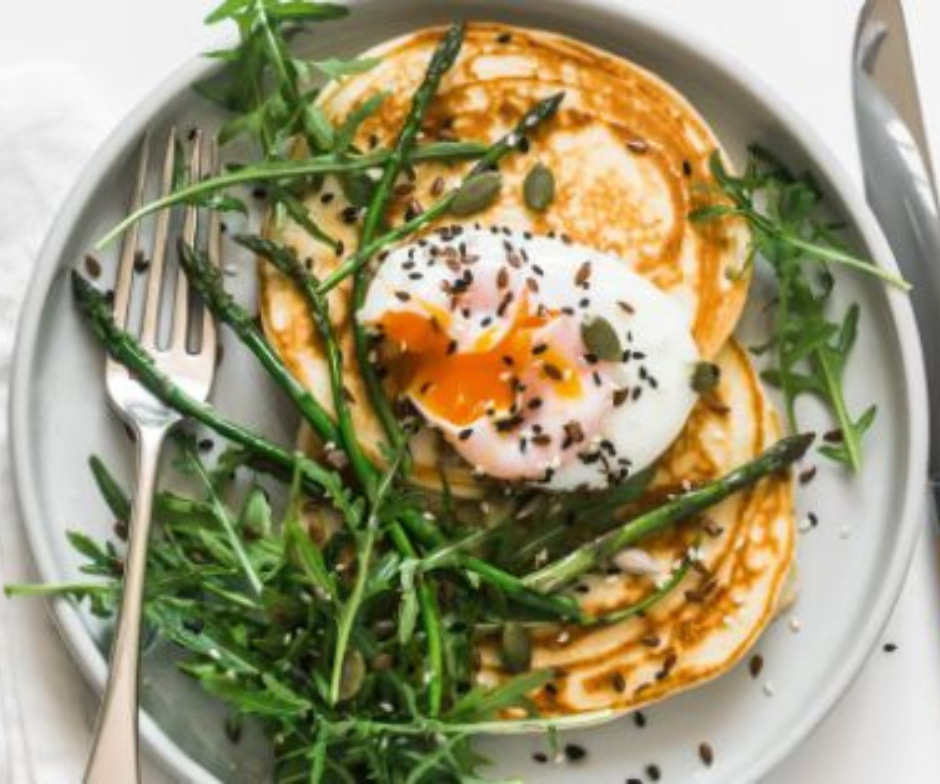Twee hartige pannenkoeken, belegd met een gepocheerd ei, asperges, rucola en zaden, geserveerd op een lichtgrijs bord met een vork.