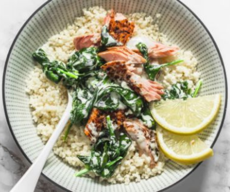 Een kom couscous met gegrilde zalm en spinazie in een romige saus, gegarneerd met twee schijfjes citroen.