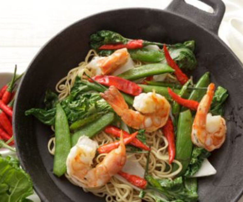 Garnalen roerbakgerecht met noedels, sugarsnaps, bladgroenten en rode chilipepers in een zwarte wok.