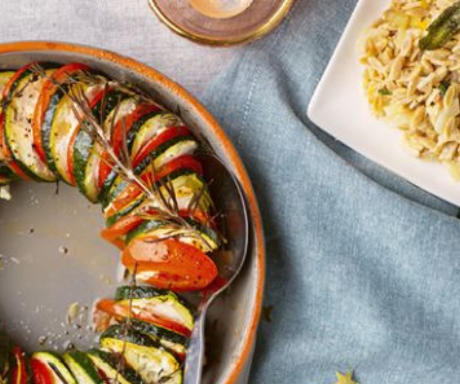 Een ring van dun gesneden geroosterde courgette en tomaten, gegarneerd met kruiden, in een kom, met een portie pasta op een blauw tafelkleed.