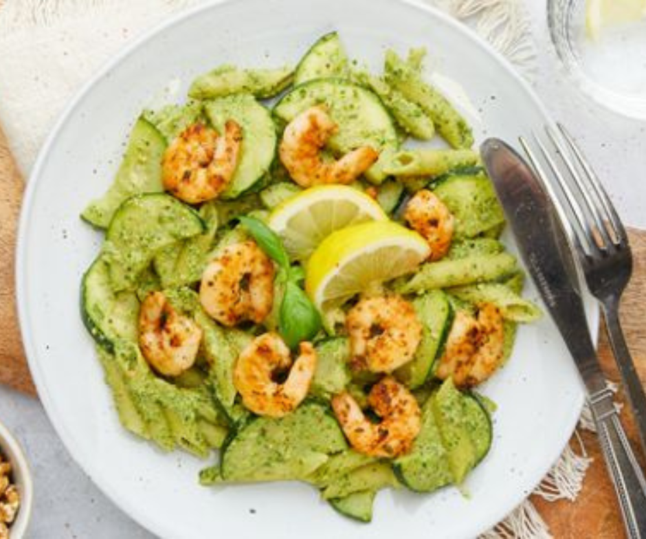 Een wit bord pasta met groene pestosaus, gebakken garnalen, plakjes komkommer en verse citroenpartjes.