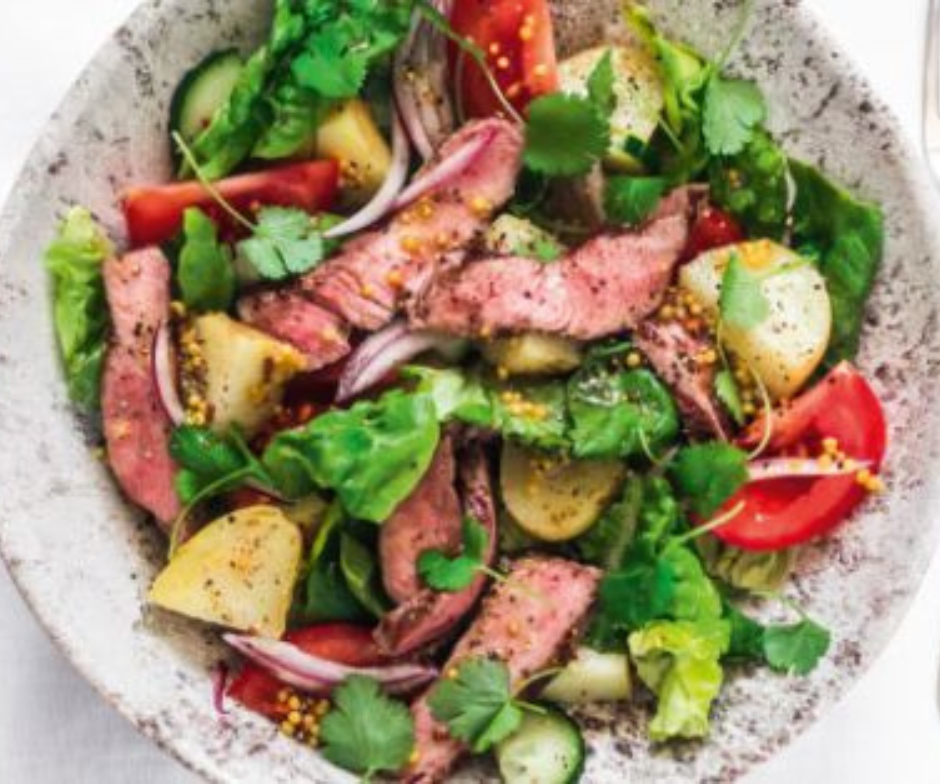 Een bovenaanzicht van een kom met een steaksalade met plakjes rundvlees, aardappelen, rode ui, tomaten en verse kruiden.