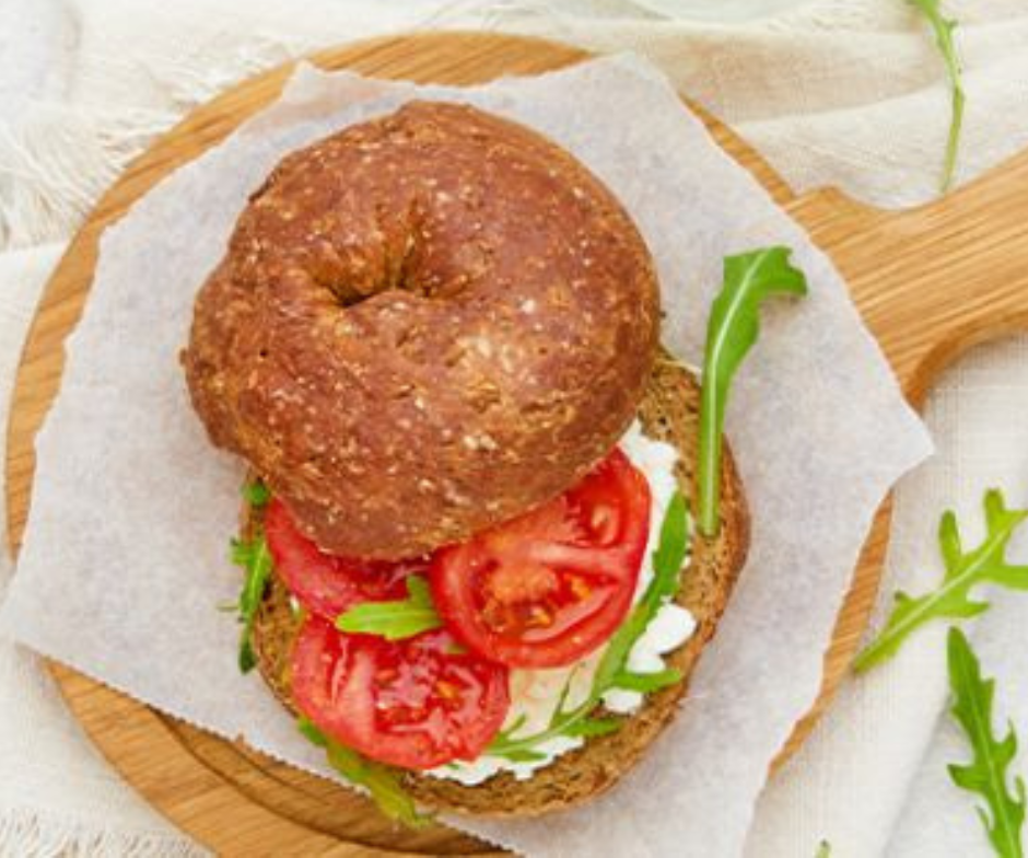 Een volkoren bagel belegd met roomkaas, plakjes tomaat en rucola, geserveerd op bakpapier op een houten plank.