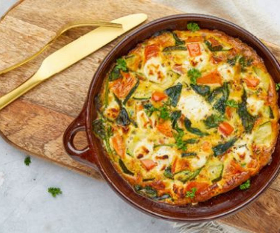 Een groentefrittata met courgette, tomaten en geitenkaas in een bruine schaal op een houten plank met een gouden mes.
