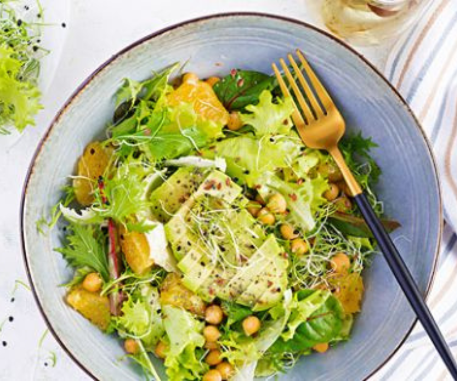 Een blauwe kom gevuld met een salade van bladgroenten, avocadoplakjes, kikkererwten en sinaasappelpartjes, met een gouden vork erin.