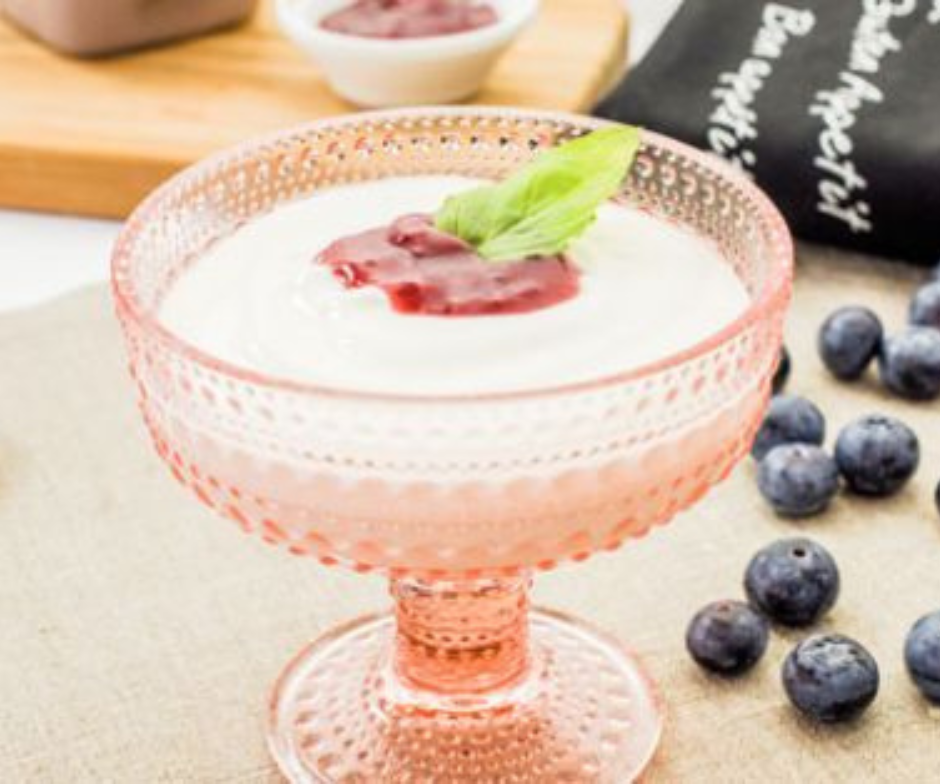 In een roze schaal op een voetstuk ligt romige witte yoghurt, gegarneerd met bessencompote en een basilicumblaadje, naast losse bosbessen.