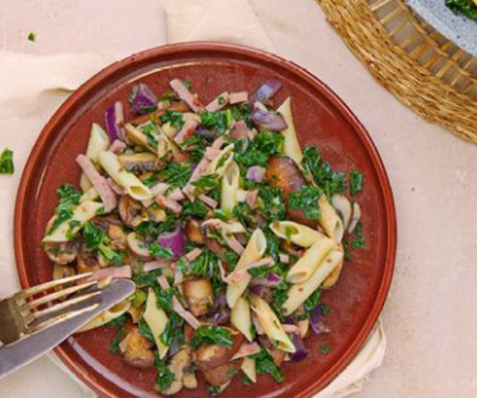 Een gerecht met penne pasta, champignons, rode ui en fijngehakte groene bladgroenten, geserveerd op een donkerrood bord met mes en vork.