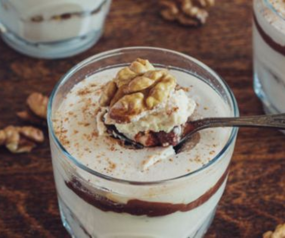 In een glazen kommetje met een romig, gelaagd dessert, gegarneerd met een walnoot, staat een lepel omhoog, op een houten tafel.