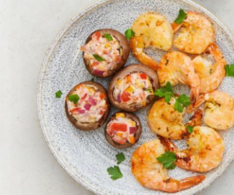 Een bord met vier gevulde champignons, gegarneerd met kruiden, en diverse gekookte garnalen op een grijze, gestructureerde achtergrond.