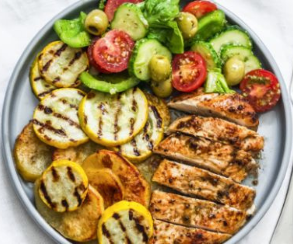 Een bord met plakjes gegrilde kip, geroosterde gele courgette en een frisse salade van tomaten, komkommers en olijven.