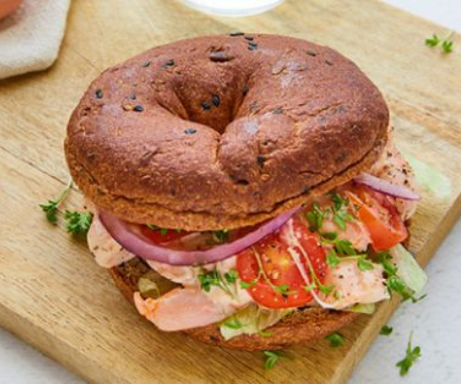 Een volkoren bagel belegd met gerookte zalm, rode ui, tomaat en verse kruiden, geserveerd op een houten snijplank.