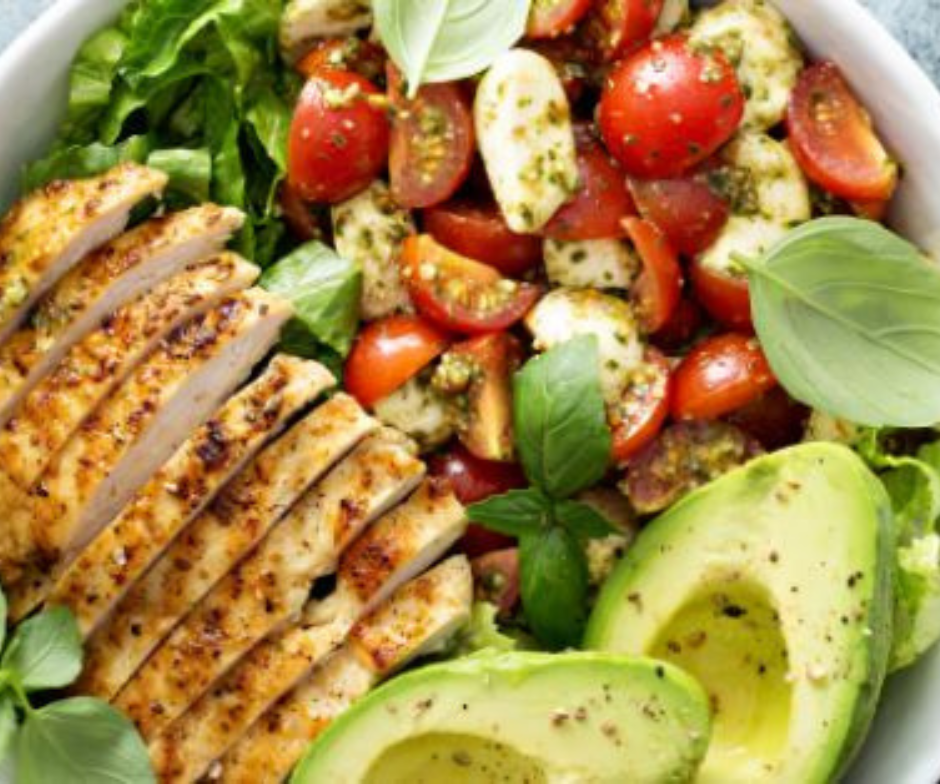Een kom met plakjes gegrilde kipfilet, een salade van tomaat en mozzarella met basilicum, en plakjes avocado op een bedje van sla.