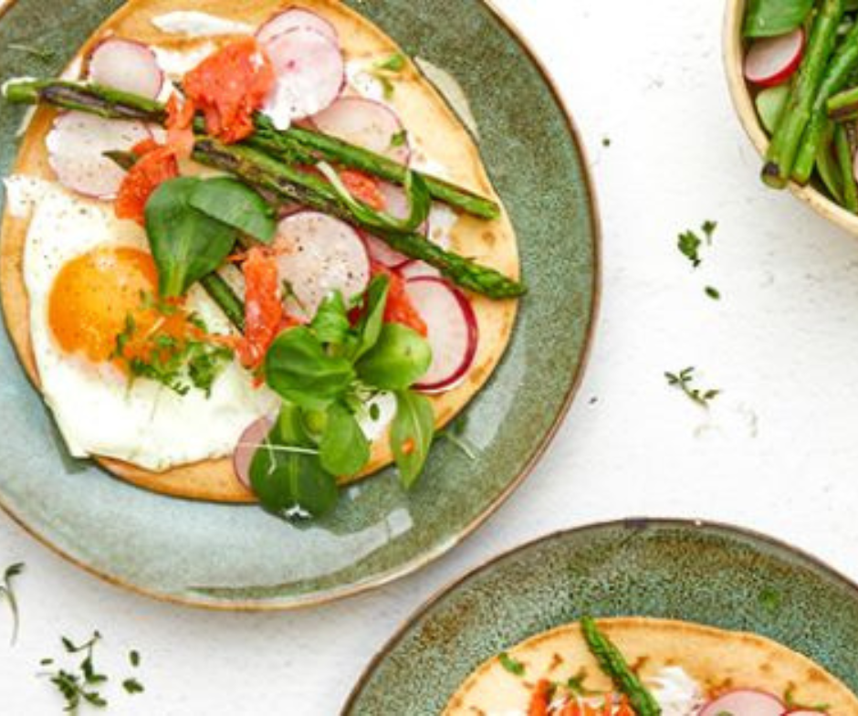 Platbroden belegd met gebakken eieren, asperges, gerookte zalm, radijsschijfjes en groene bladgroenten, geserveerd op groene keramische borden.