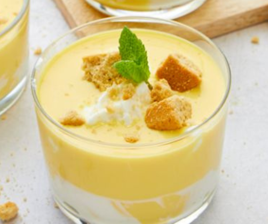 Een gelaagd dessert in een glas met witte room, gele pudding, koekjeskruimels en een takje munt als garnering.