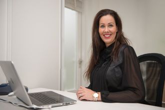 Een professionele vrouw met lang donker haar zit met een laptop achter een bureau in een lichte kantoorruimte en glimlacht.