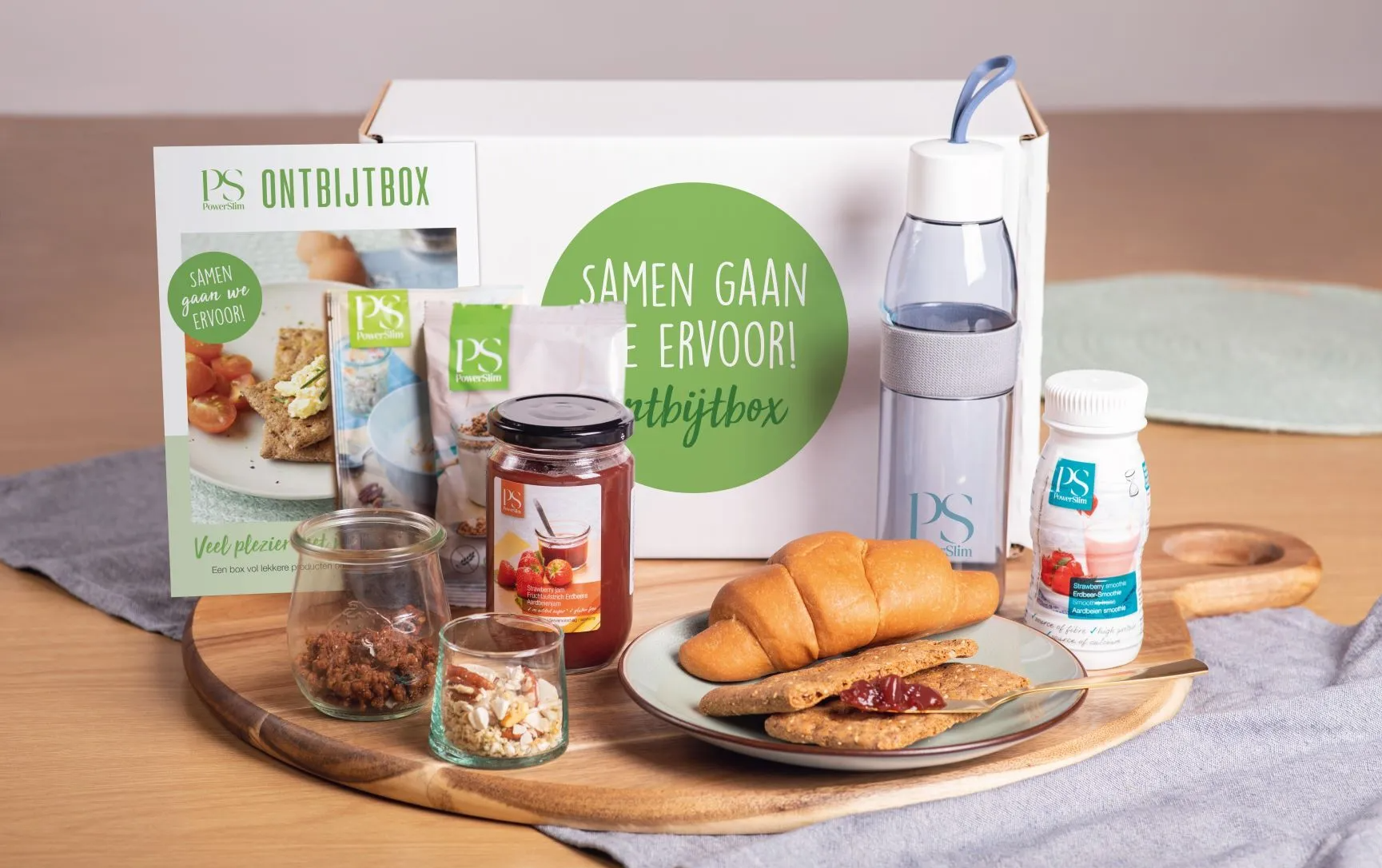 Een ontbijtbox met snacks, jam, een flesje drinken en een gebakje, uitgestald op een houten plank op tafel.