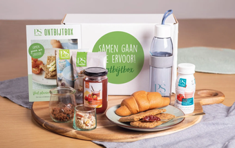 Een promotionele ontbijtbox met etenswaren, een herbruikbare waterfles en informatiemateriaal op een houten plank.