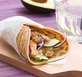 Een kipwrap met courgette en rode ui op een houten plank, met een avocado en een glas water ernaast.