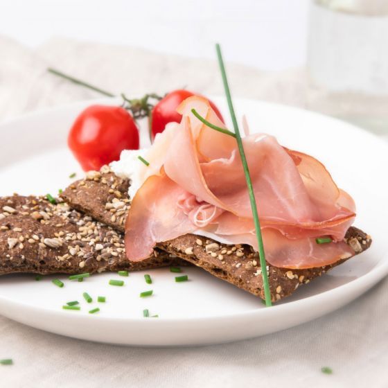 Een sneetje knäckebröd belegd met roomkaas en gevouwen ham, gegarneerd met bieslook en cherrytomaatjes, op een wit bord.