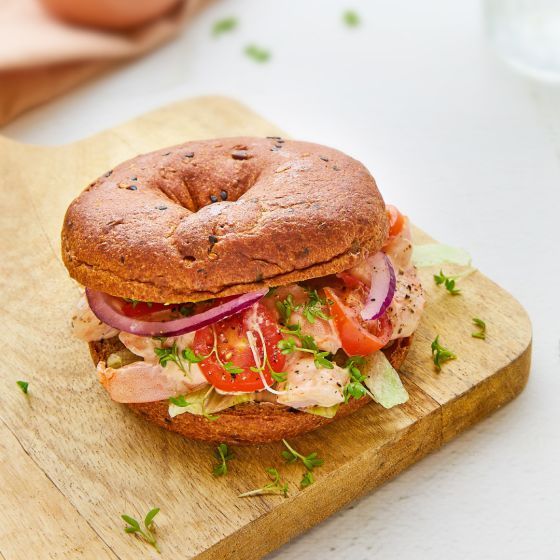 Een bagelsandwich met zalm, plakjes tomaat, rode ui en verse groene bladgroenten, geserveerd op een houten plank.