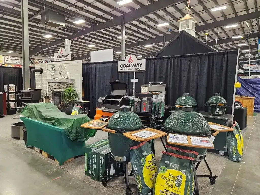 Display of grills and grilling accessories at a trade show booth, with green egg smokers and black backdrop.