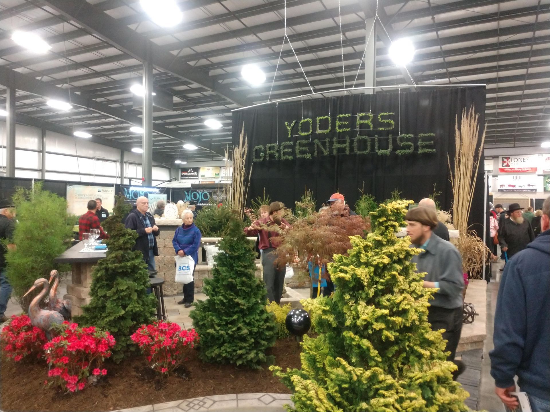 Yoder's Greenhouse booth at an event. Display of plants with people gathered. Dark sign reads YODERS GREENHOUSE.