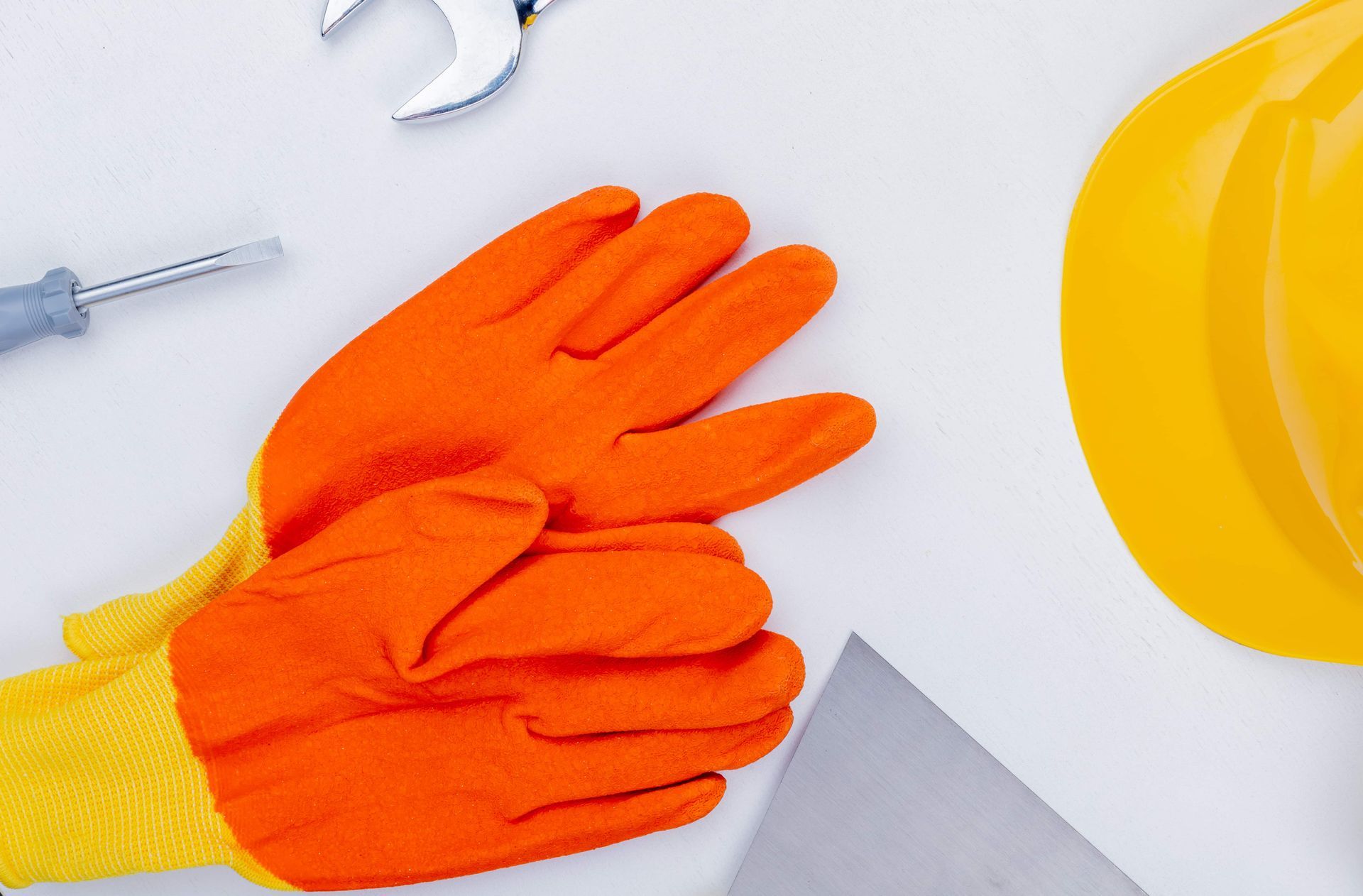 Orange work gloves beside a yellow hard hat and gray tool on a white surface