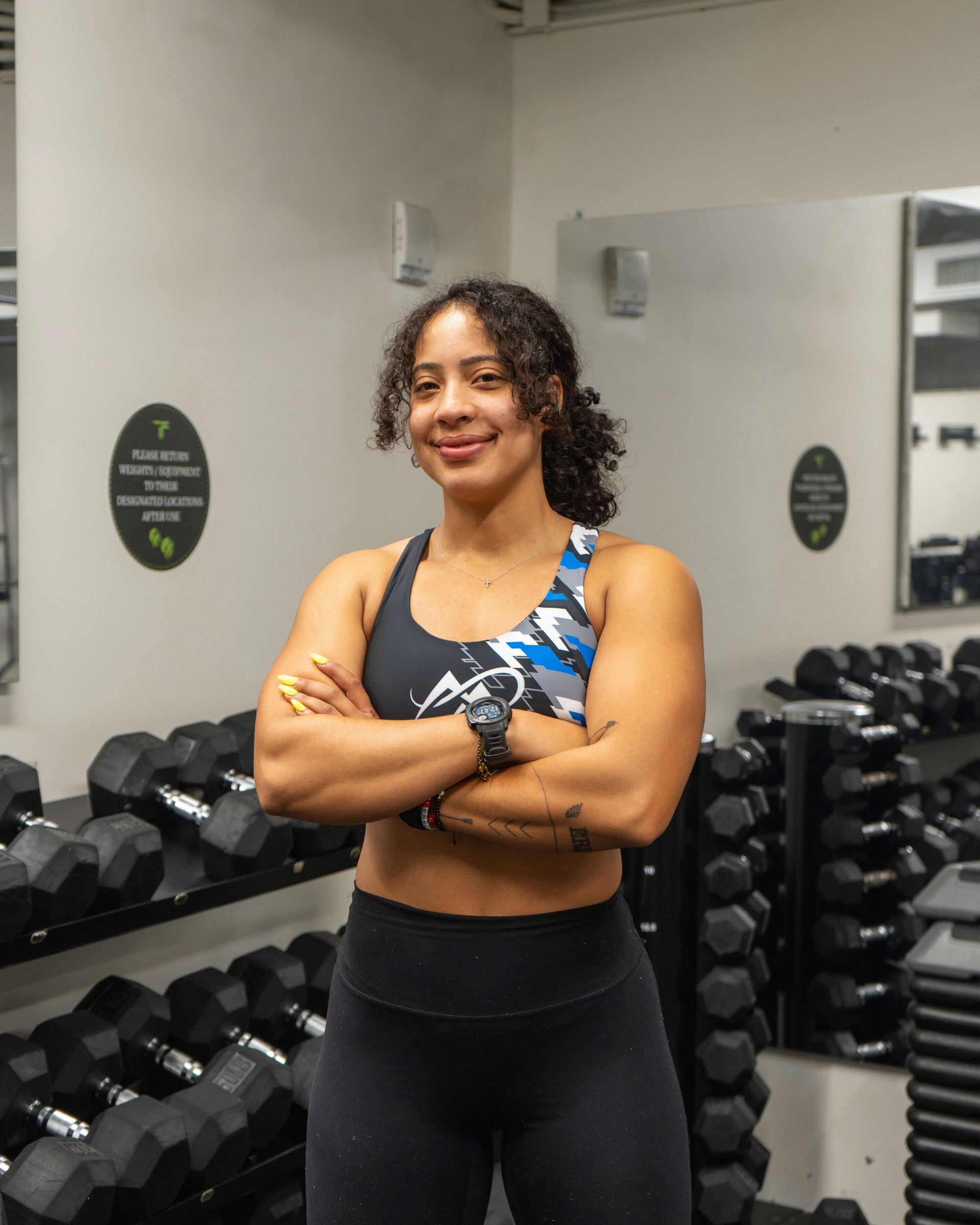 Woman in workout clothes, arms crossed, standing in a gym with weights.