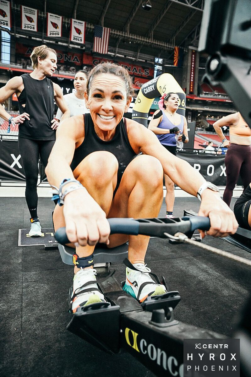 Athlete rowing in a HYROX race, smiling, on a black indoor competition floor.