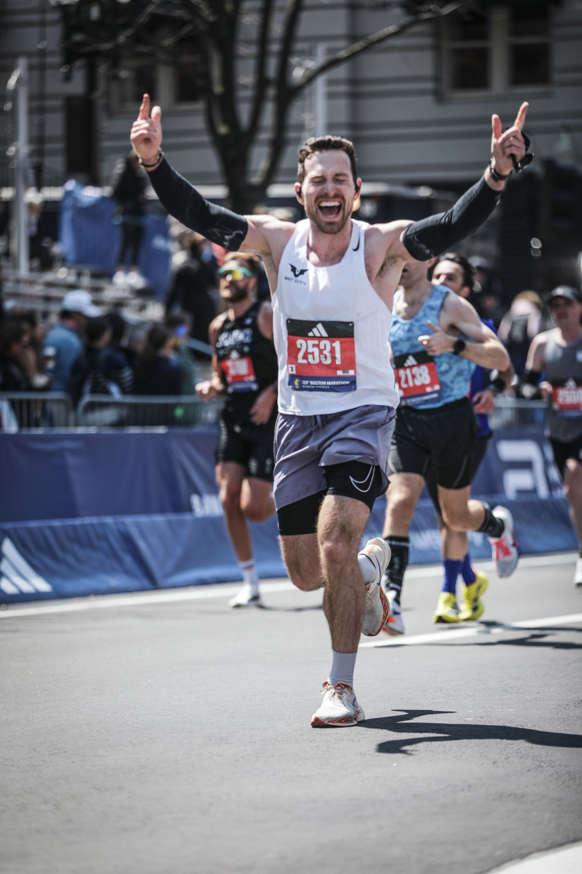 Runner with arms raised, crossing finish line. He's wearing a race bib, tank top, and shorts.