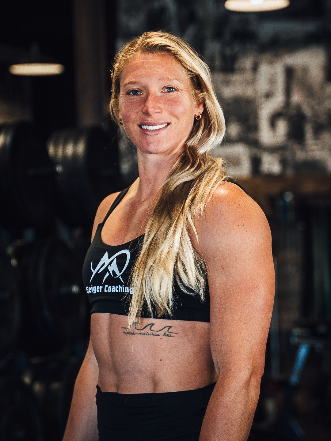 Woman with blonde hair and muscular arms in a gym, smiling, wearing a sports bra and shorts.