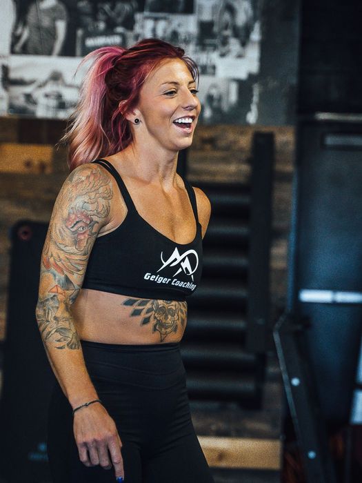 Woman with pink hair laughs, wearing a black sports bra and leggings, in a gym.