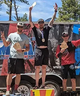 Three people stand on a podium, holding awards. Middle person raises arms, celebrating. Others smile, posing.
