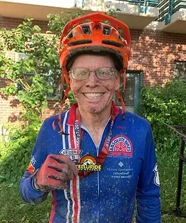 Man in orange helmet and blue cycling jersey, covered in mud, holding a medal, smiling.
