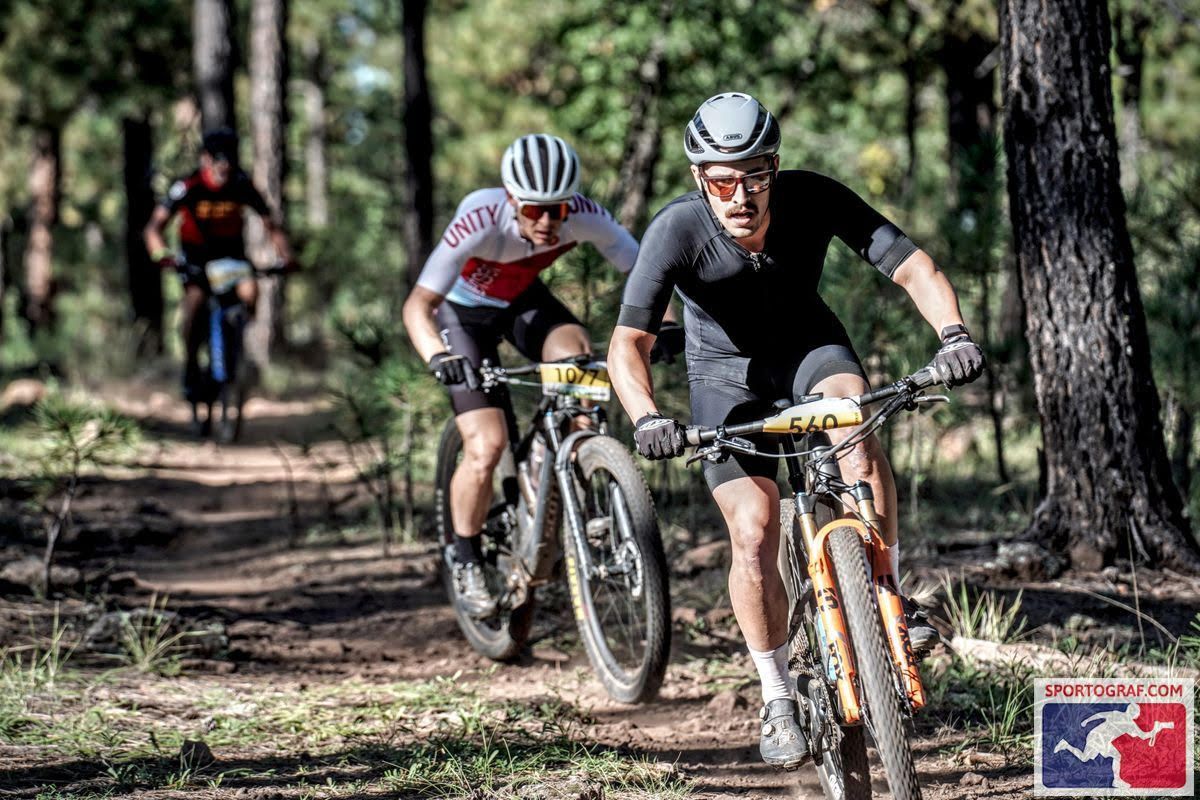 Mountain bikers racing on a dirt trail in a forest, bright sunlight, wearing helmets and athletic gear.