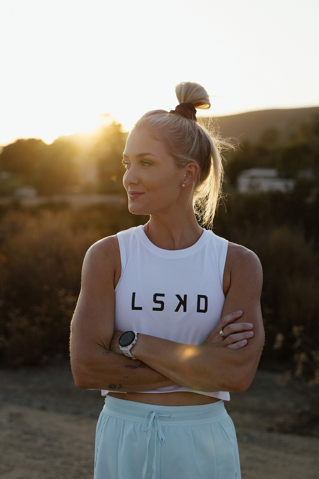 Woman with arms crossed, wearing white tank top with 'LSKD' logo, in sunlight.