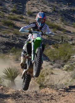 Dirt bike rider airborne on a green and white motorcycle in a desert landscape.