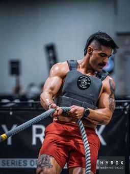 Man in weighted vest pulls battle rope, intense expression, red shorts, indoor gym.