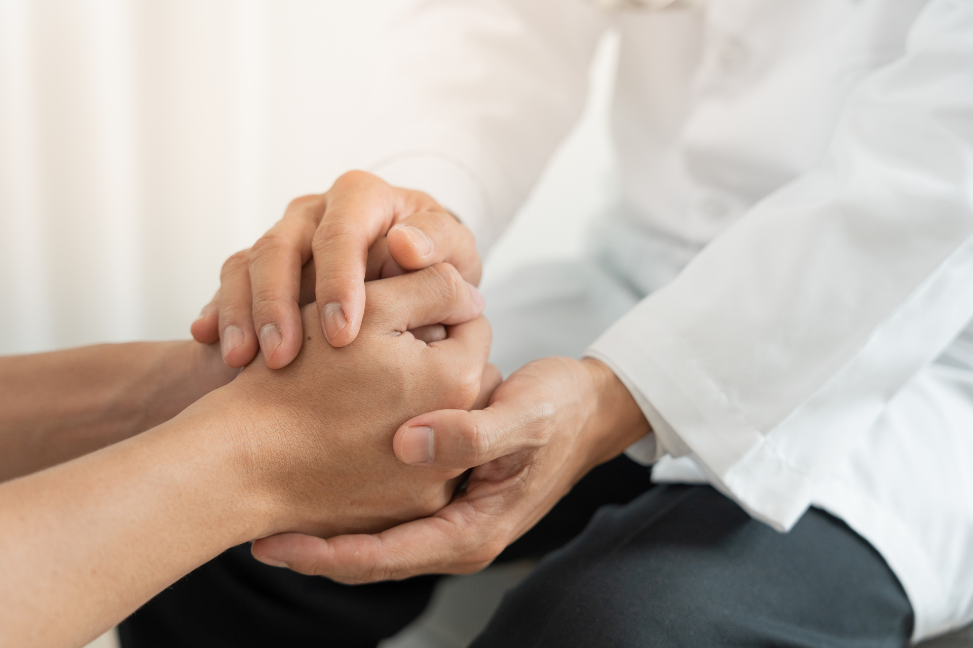 A person in a white medical coat gently holds the hands of another person, showing comfort and support.