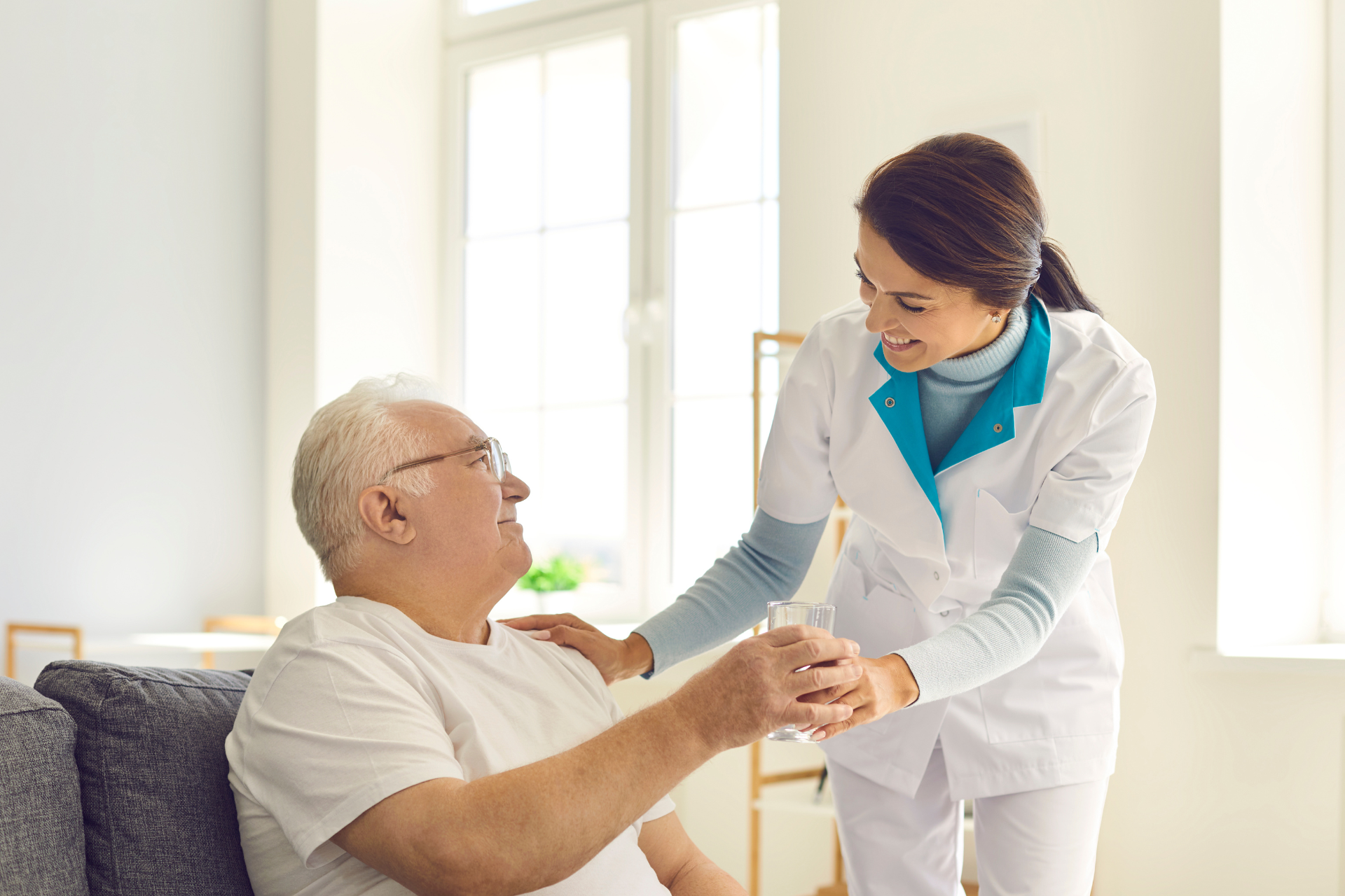 A professional caregiver in a white uniform hands a glass of water to an older person sitting on a sofa in a bright room.