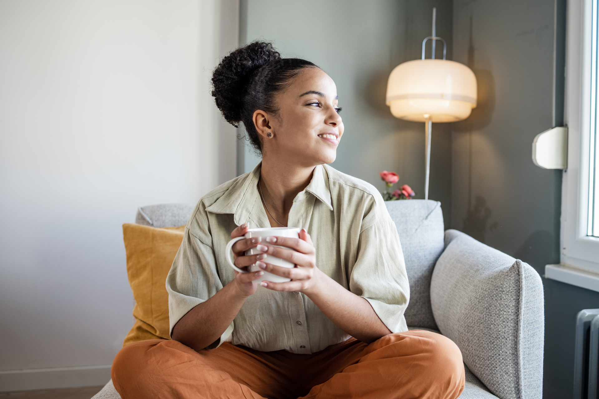 A person with hair in a bun sits on a couch holding a white mug, looking toward a window next to a floor lamp.
