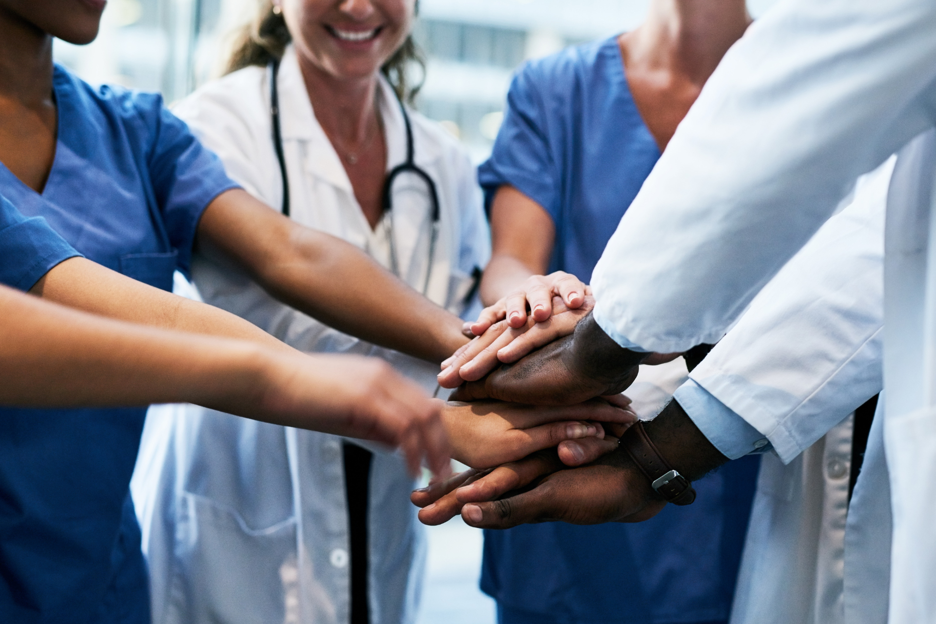 A group of healthcare workers in scrubs and lab coats stacking their hands together in a symbol of teamwork.