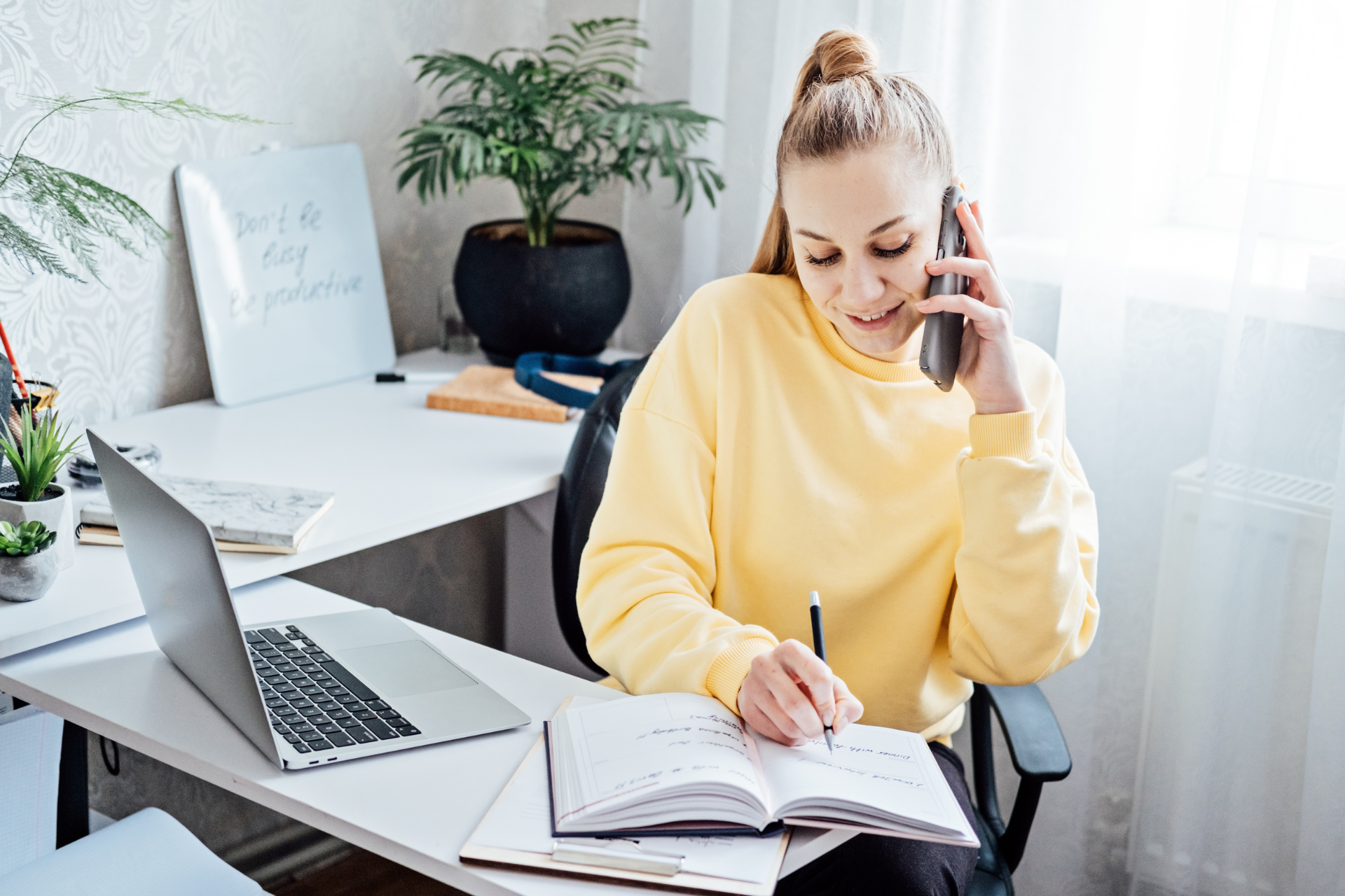 A person in a yellow sweatshirt sits at a desk, talking on a smartphone and writing in a notebook near a laptop.