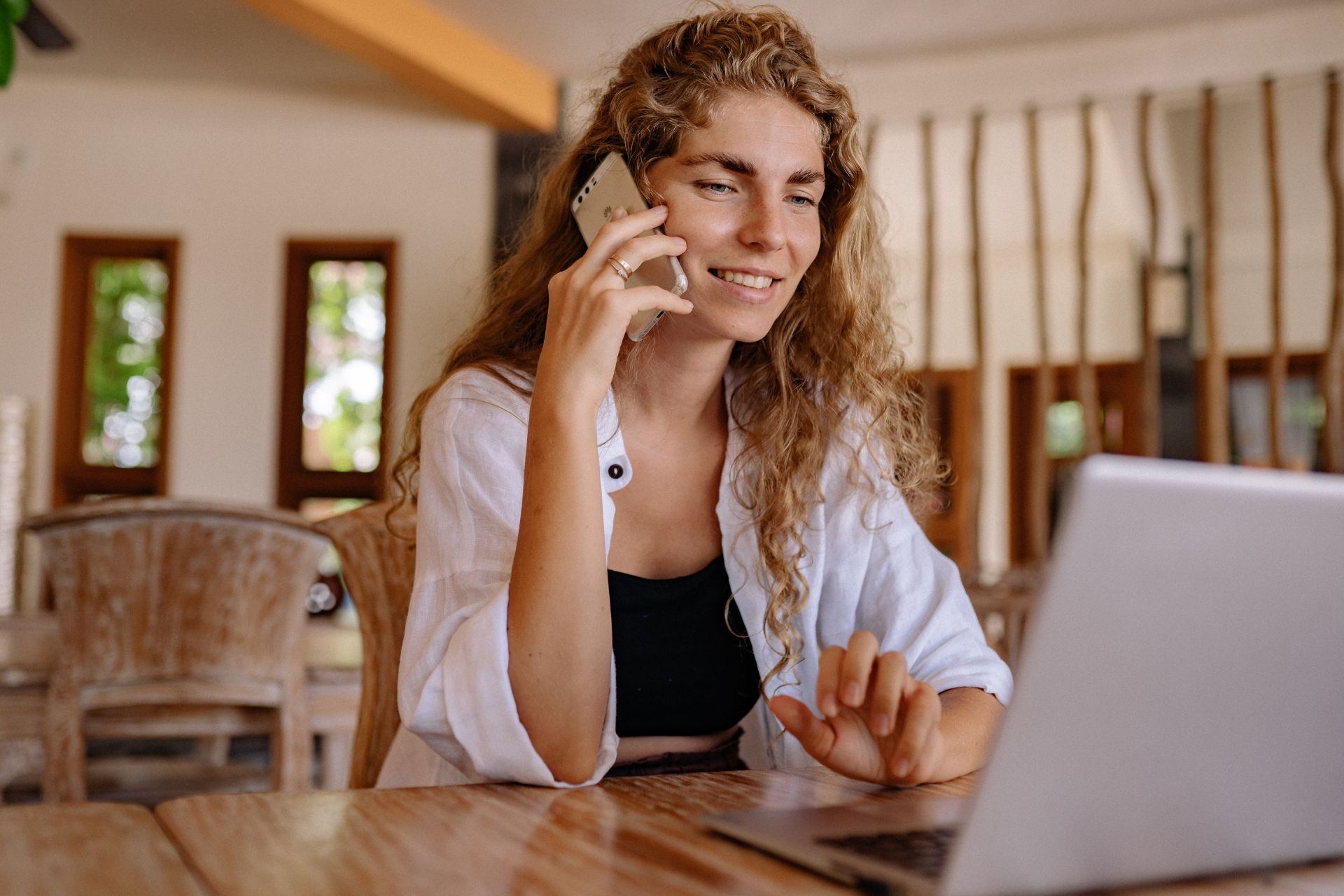 A person with curly hair talking on a smartphone while sitting at a table with a laptop in a bright, rustic setting.