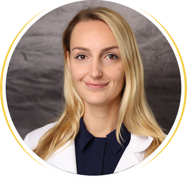 Headshot of a person with blonde hair smiling, wearing a white medical coat and navy shirt against a gray background.